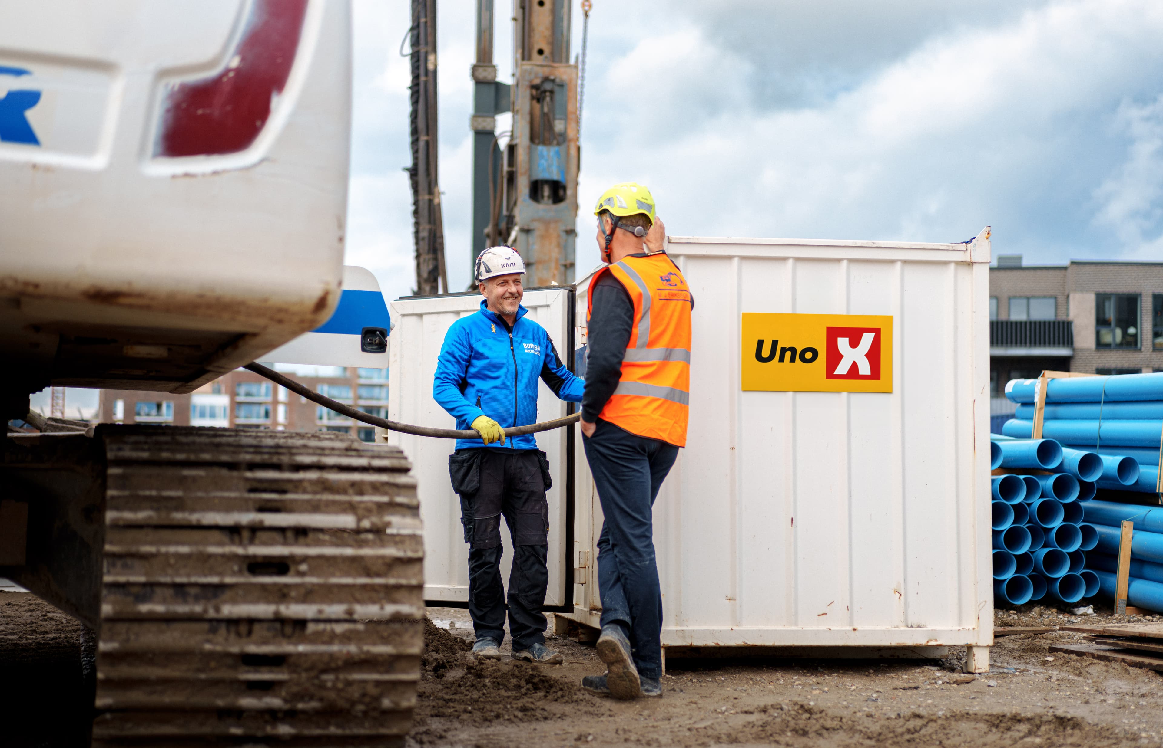 To mænd ved en Uno X brændstofcontainer på en byggeplads med maskiner og materialer, der illustrerer praktisk brændstofhåndtering.